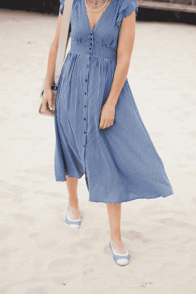 A woman in a blue gingham summer dress walking along a Nantucket beach shore.