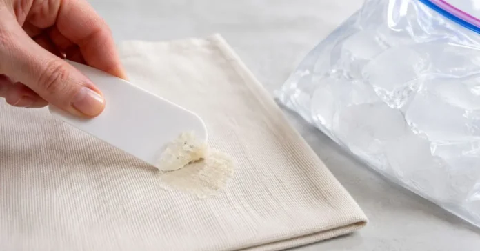 Hardened hot glue being lifted from cotton fabric with a plastic scraper beside a sealed ice bag